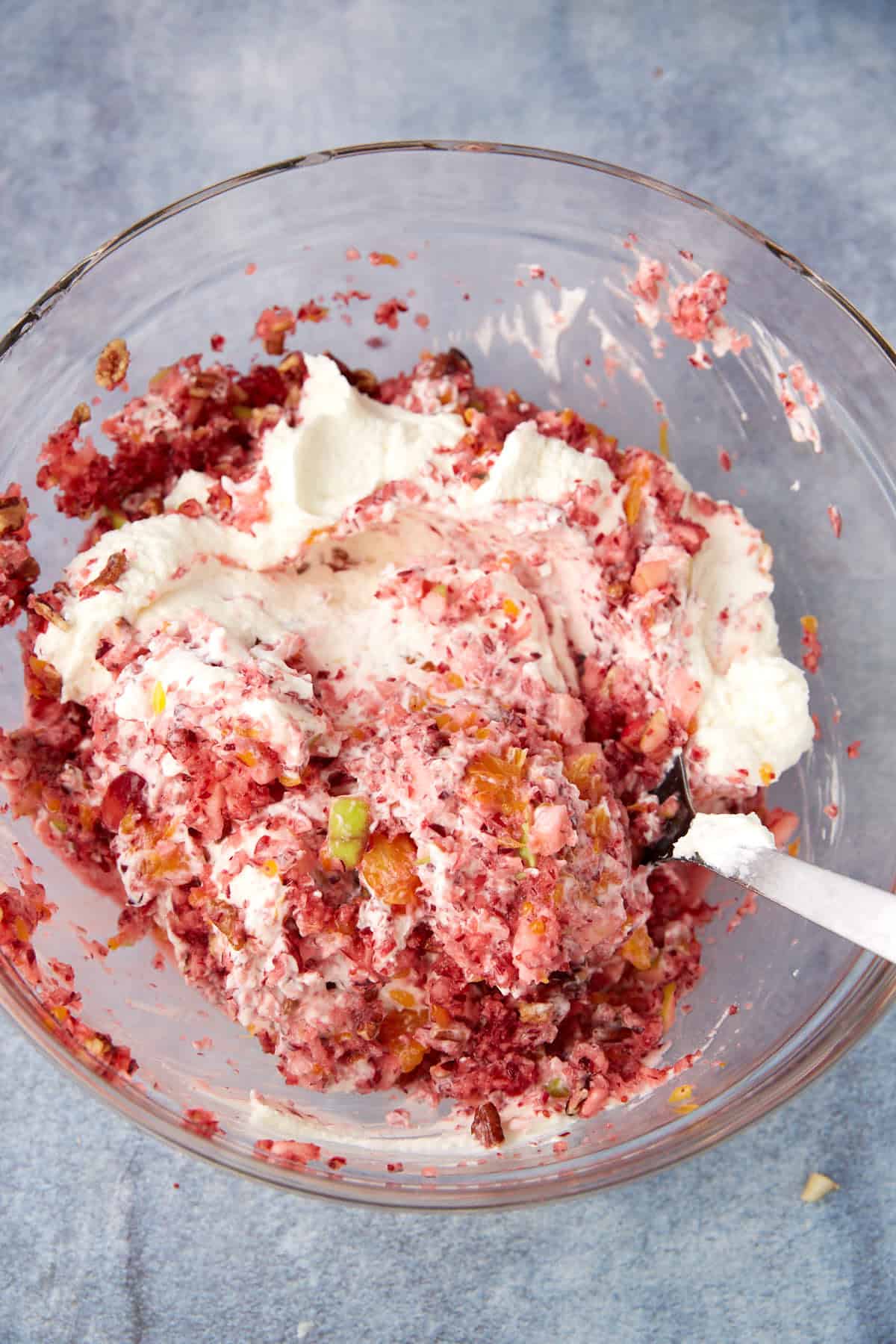 A glass bowl containing a mixture of chopped red fruit, green and yellow bits, and whipped cream, with a spoon partially buried in the mixture on a blue-gray surface.