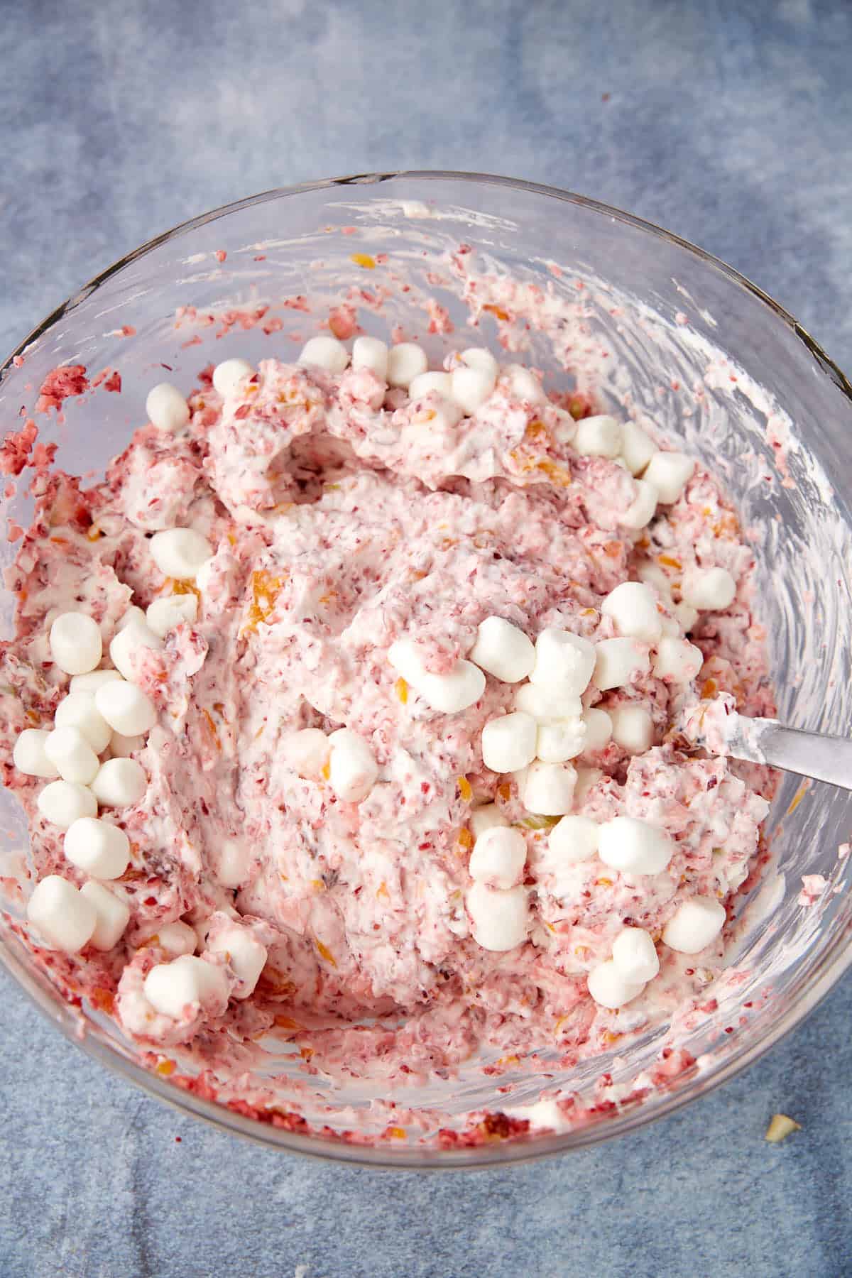 A glass bowl filled with a pink, creamy mixture containing bits of fruit and small white mini marshmallows, with a spoon resting inside. The bowl is placed on a blue surface.