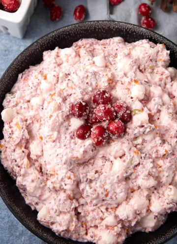 A black bowl filled with creamy pink salad, mixed with marshmallows and fruit, garnished with sugared cranberries on top. A small white container of cranberries sits in the background.