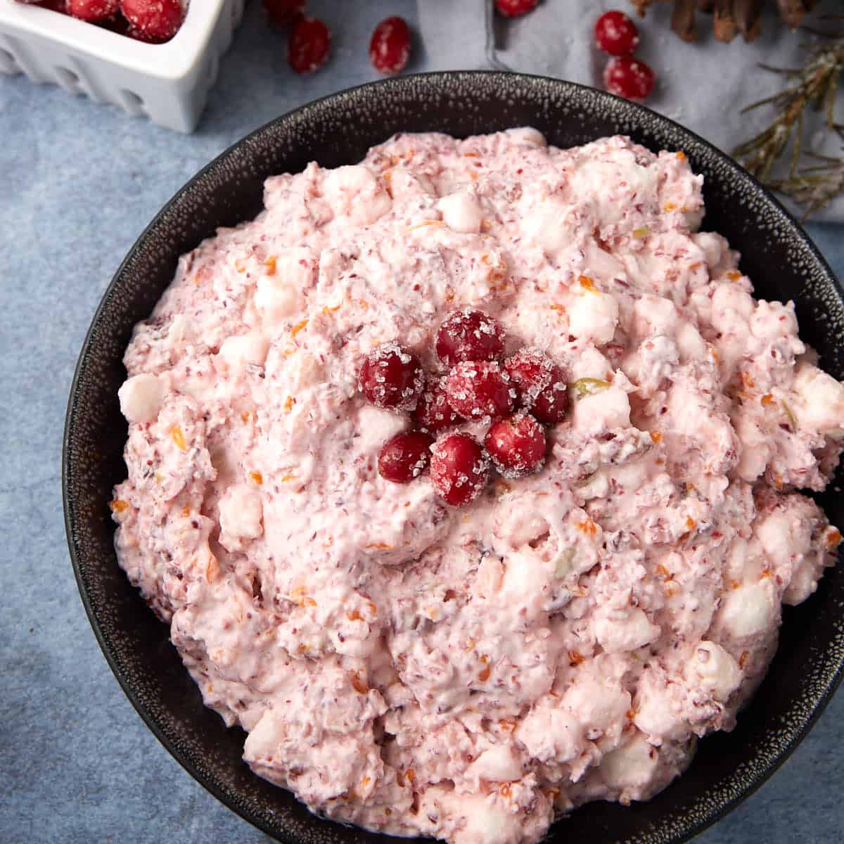 A black bowl filled with creamy pink salad, mixed with marshmallows and fruit, garnished with sugared cranberries on top. A small white container of cranberries sits in the background.