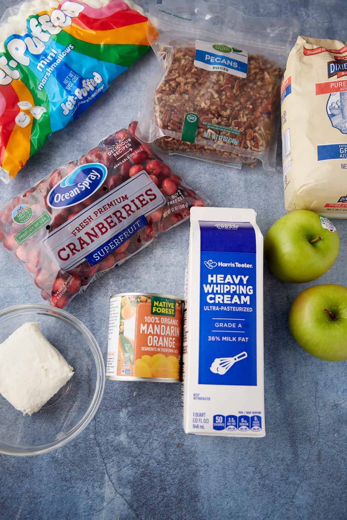 Various ingredients on a blue surface: mini marshmallows, chopped pecans, flour, fresh cranberries, a can of mandarin oranges, two green apples, heavy whipping cream, and a small bowl of cream cheese.