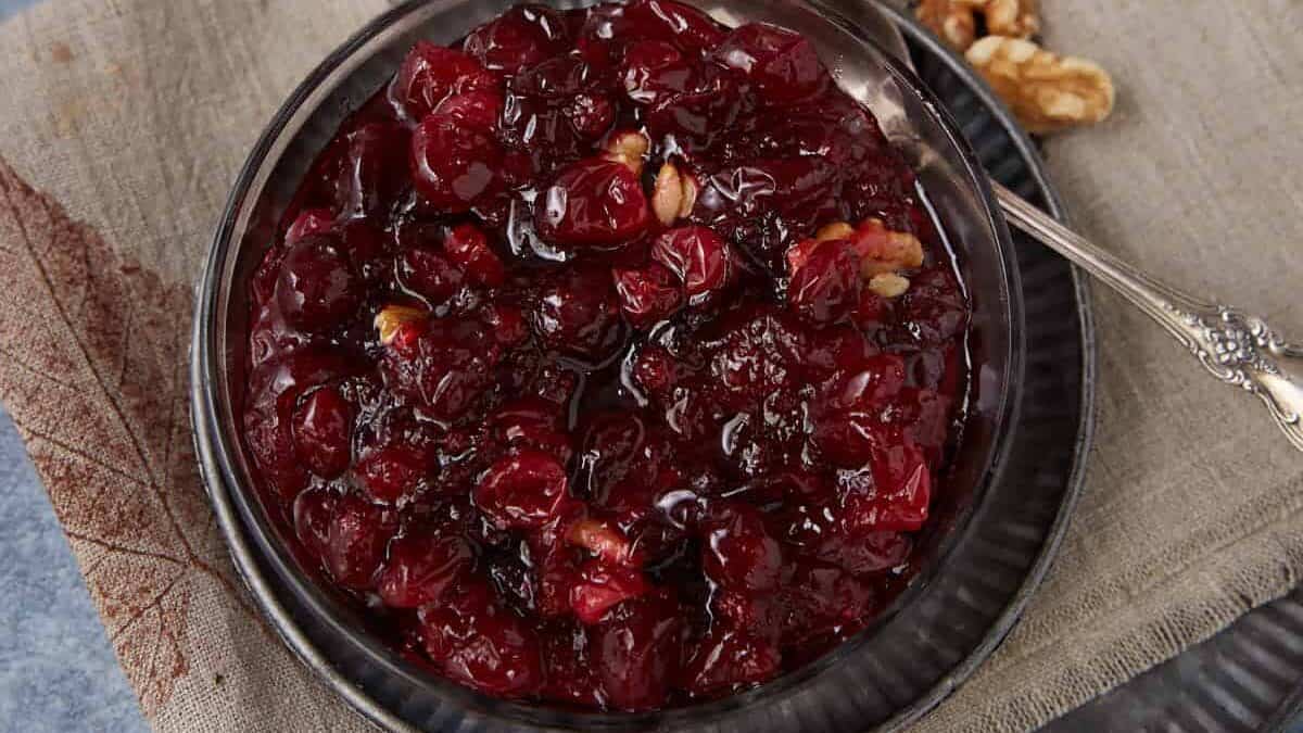 A bowl of homemade cranberry sauce with walnuts sits on a beige napkin on a dark plate, with a silver spoon and extra walnut pieces beside it on a blue and gray background.