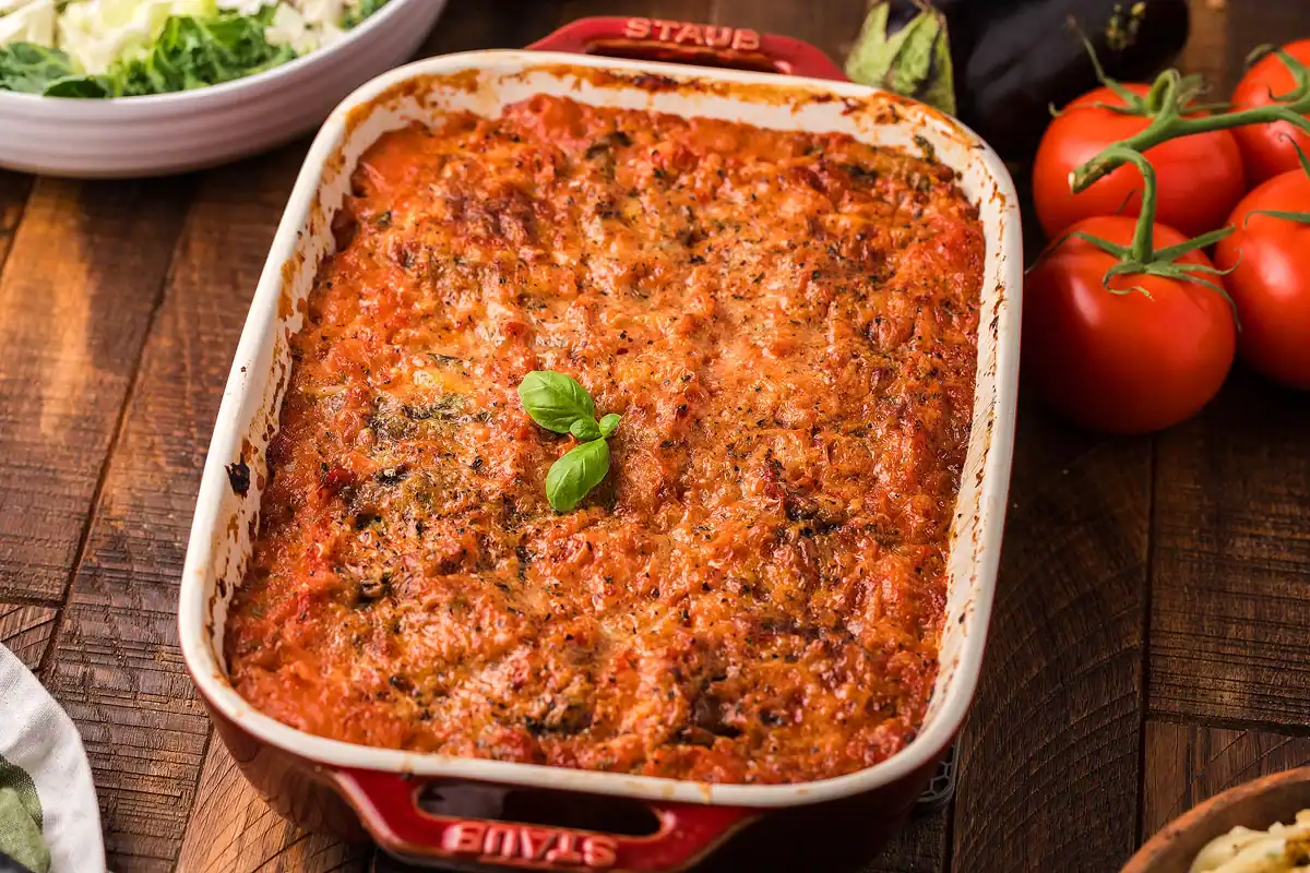A freshly baked dish of lasagna topped with melted, golden cheese and garnished with fresh basil, in a red baking dish on a wooden table with tomatoes and lettuce nearby.
