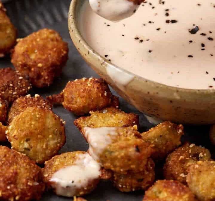 A hand dips a piece of fried okra into a bowl of creamy dipping sauce on a plate filled with crispy okra bites. The sauce is sprinkled with black pepper.