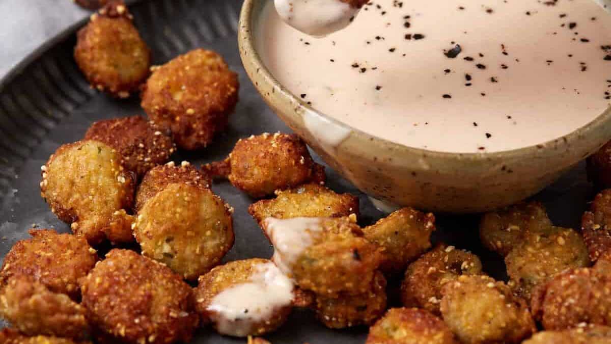 A hand dips a piece of fried okra into a bowl of creamy dipping sauce on a plate filled with crispy okra bites. The sauce is sprinkled with black pepper.