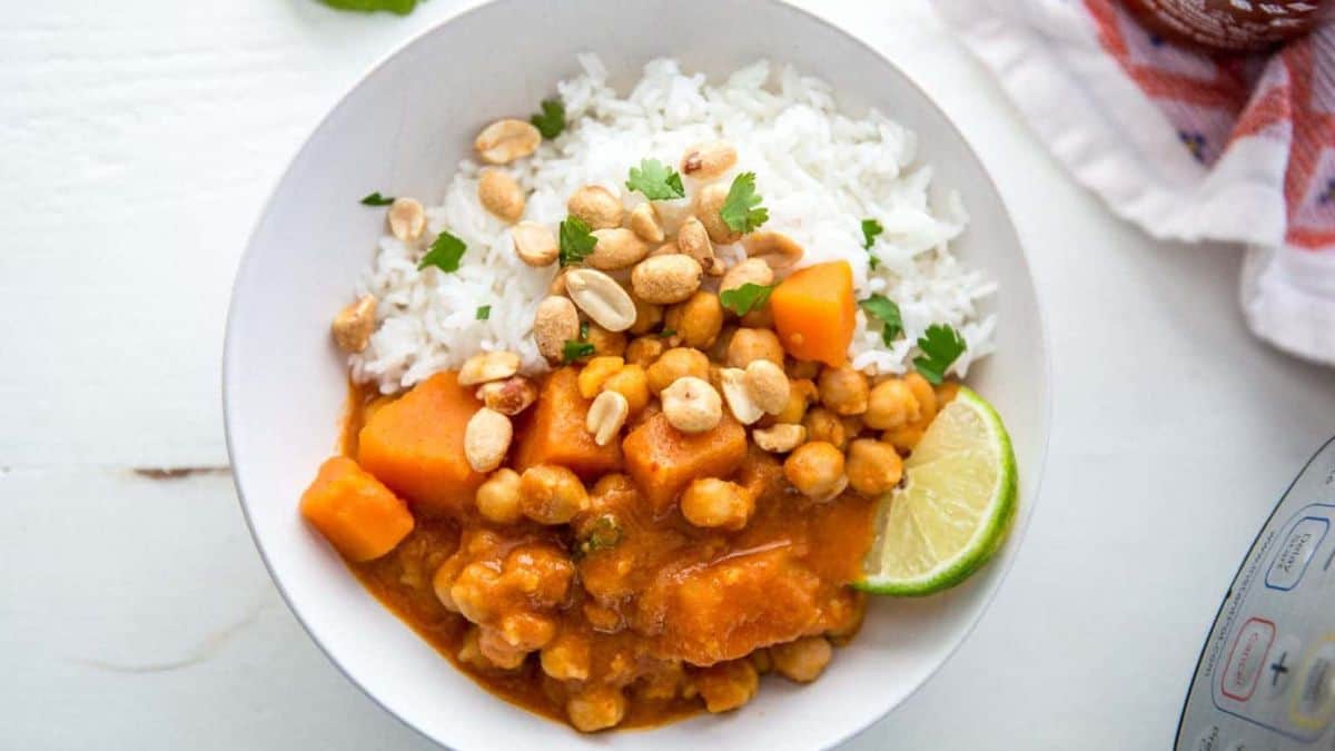 A white bowl filled with white rice and a chickpea and vegetable curry, garnished with fresh cilantro, peanuts, and a lime wedge, sits on a white surface.