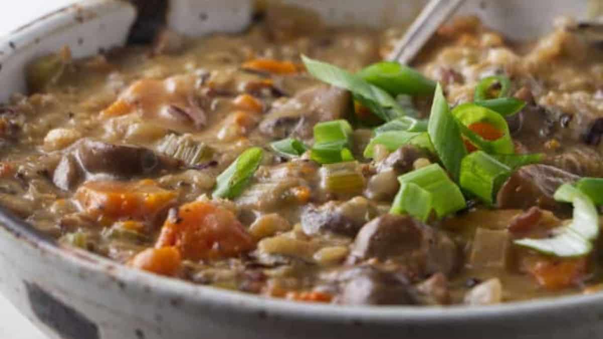 A close-up of a bowl of hearty soup with visible chunks of carrots, mushrooms, and grains, garnished with chopped green onions, and a spoon resting in the bowl.