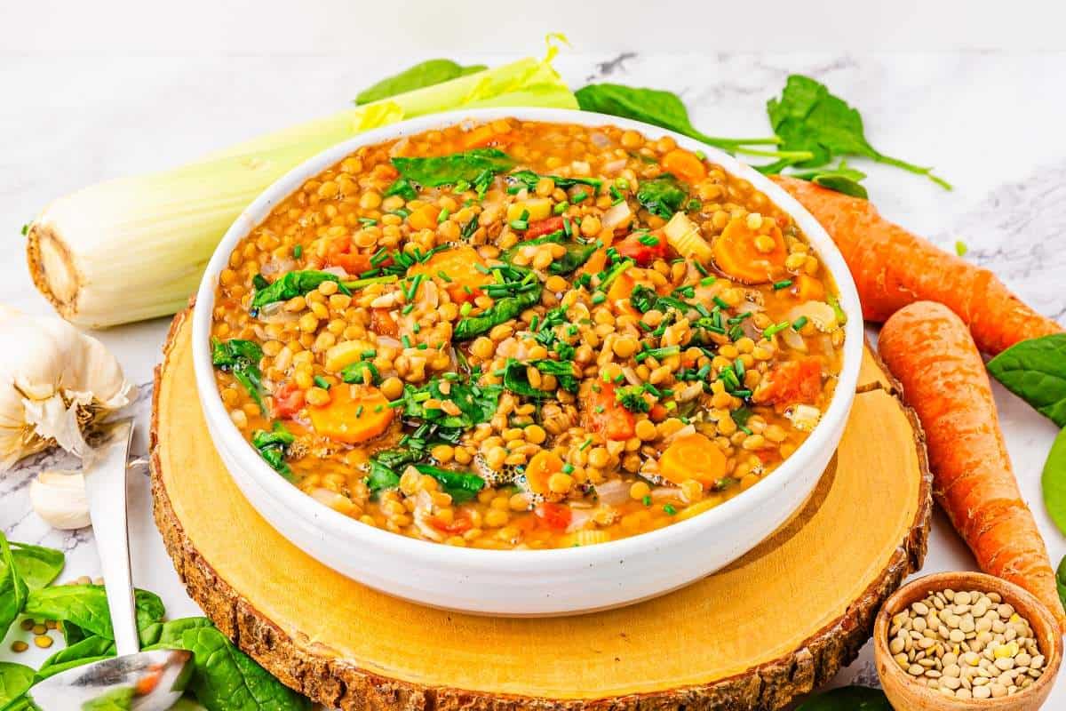 A bowl of lentil soup with carrots, spinach, and chopped herbs sits on a wooden board. Fresh carrots, spinach leaves, garlic, and dry lentils are arranged around the bowl.