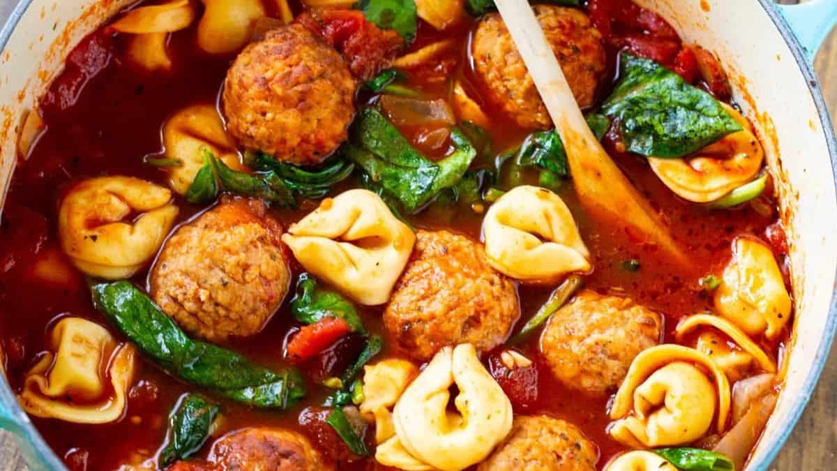A pot filled with tomato-based soup containing meatballs, cheese tortellini, spinach leaves, and diced tomatoes, with a wooden spoon resting inside.