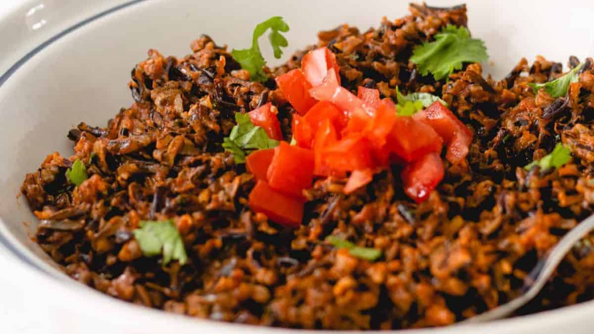 A close-up of a bowl of wild rice topped with chopped tomatoes and cilantro, with a spoon visible on the side. The rice appears seasoned and garnished for serving.