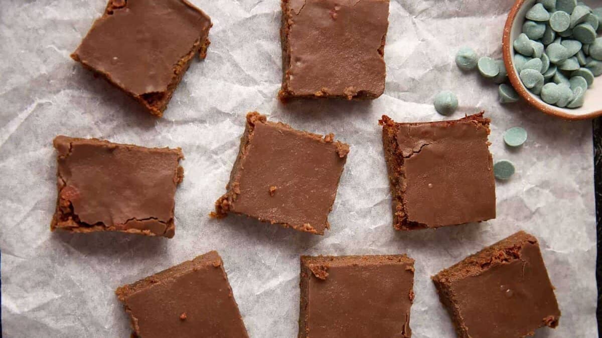 Eight chocolate-topped squares of brownie on parchment paper, with a small dish of greenish chocolate chips in the upper right corner.