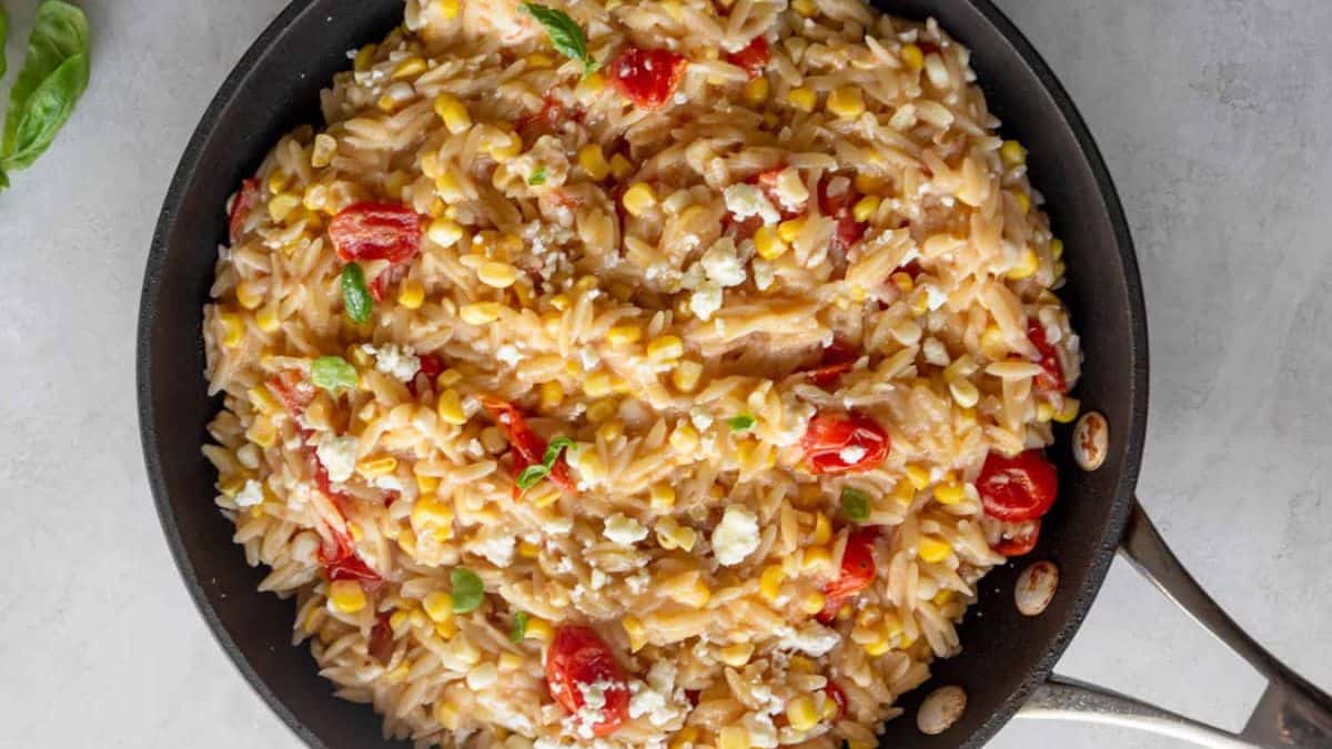 A skillet filled with creamy orzo pasta mixed with corn, cherry tomatoes, and topped with crumbled cheese and fresh herbs.