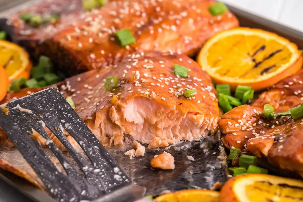 Baked salmon fillets topped with sesame seeds and chopped green onions, surrounded by orange slices on a tray—perfect for dinners under 30 minutes worth saving. A spatula lifts a piece, revealing the tender, flaky texture of the fish.