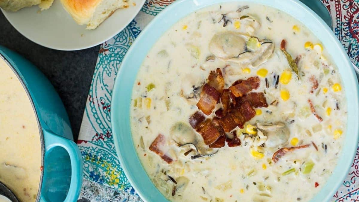 A bowl of creamy chowder topped with crispy bacon pieces, containing oysters, corn, and herbs, sits on a colorful patterned cloth next to a pot of more chowder and a plate with a bread roll.