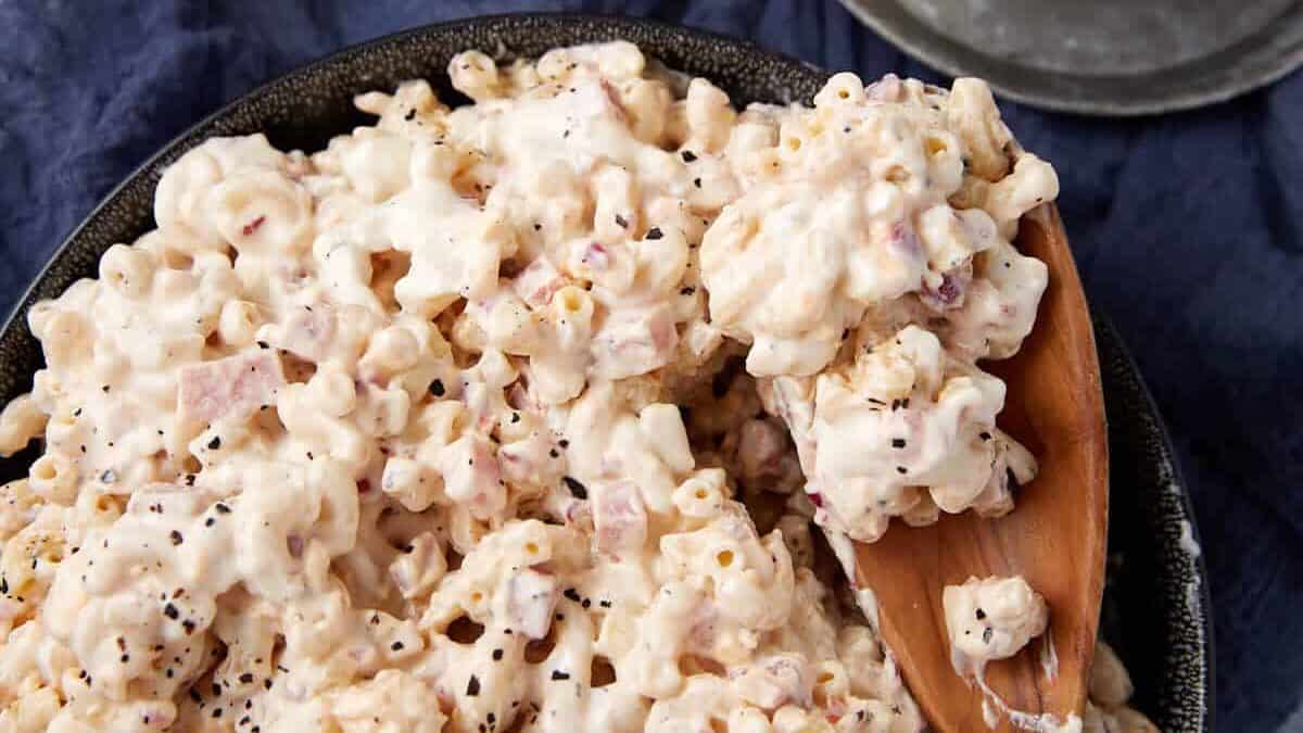 A bowl of creamy macaroni salad with a wooden spoon rests on a dark cloth, next to a small pitcher of extra dressing on a saucer.