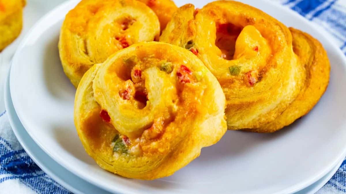 Three golden, spiral-shaped pinwheel pastries with visible bits of red and green peppers, and melted cheese, served on a white plate with a blue and white cloth in the background.