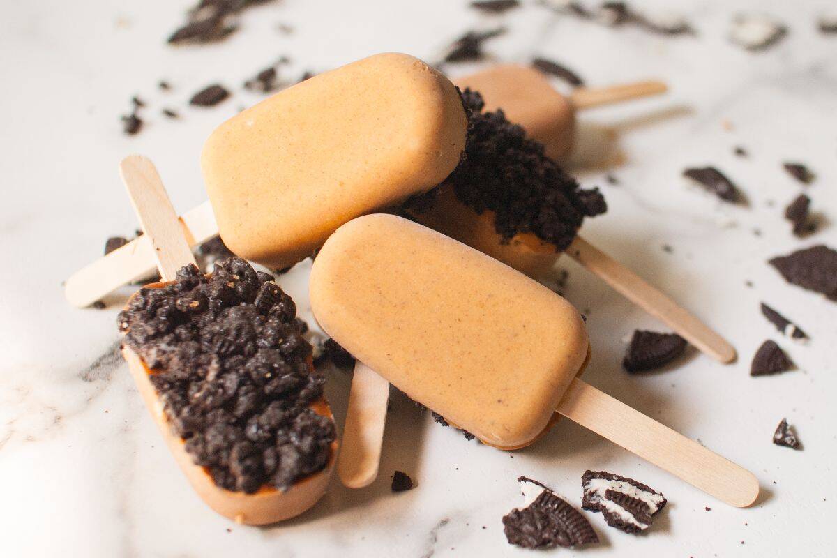 Four caramel-colored ice cream bars on wooden sticks, with two bars partially coated in crushed chocolate cookies, are arranged on a white marble surface scattered with cookie crumbs.