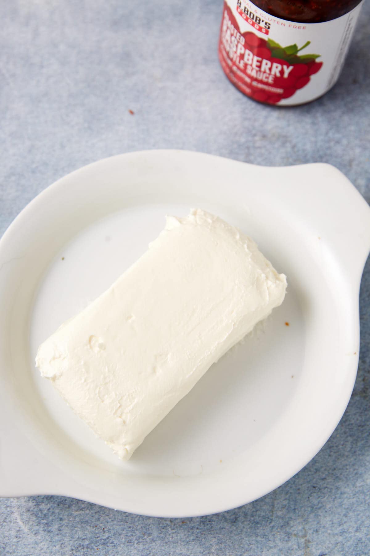 A block of cream cheese on a white plate, perfect for making a delicious raspberry cream cheese dip, with a jar of raspberry preserves partially visible in the top right corner.
