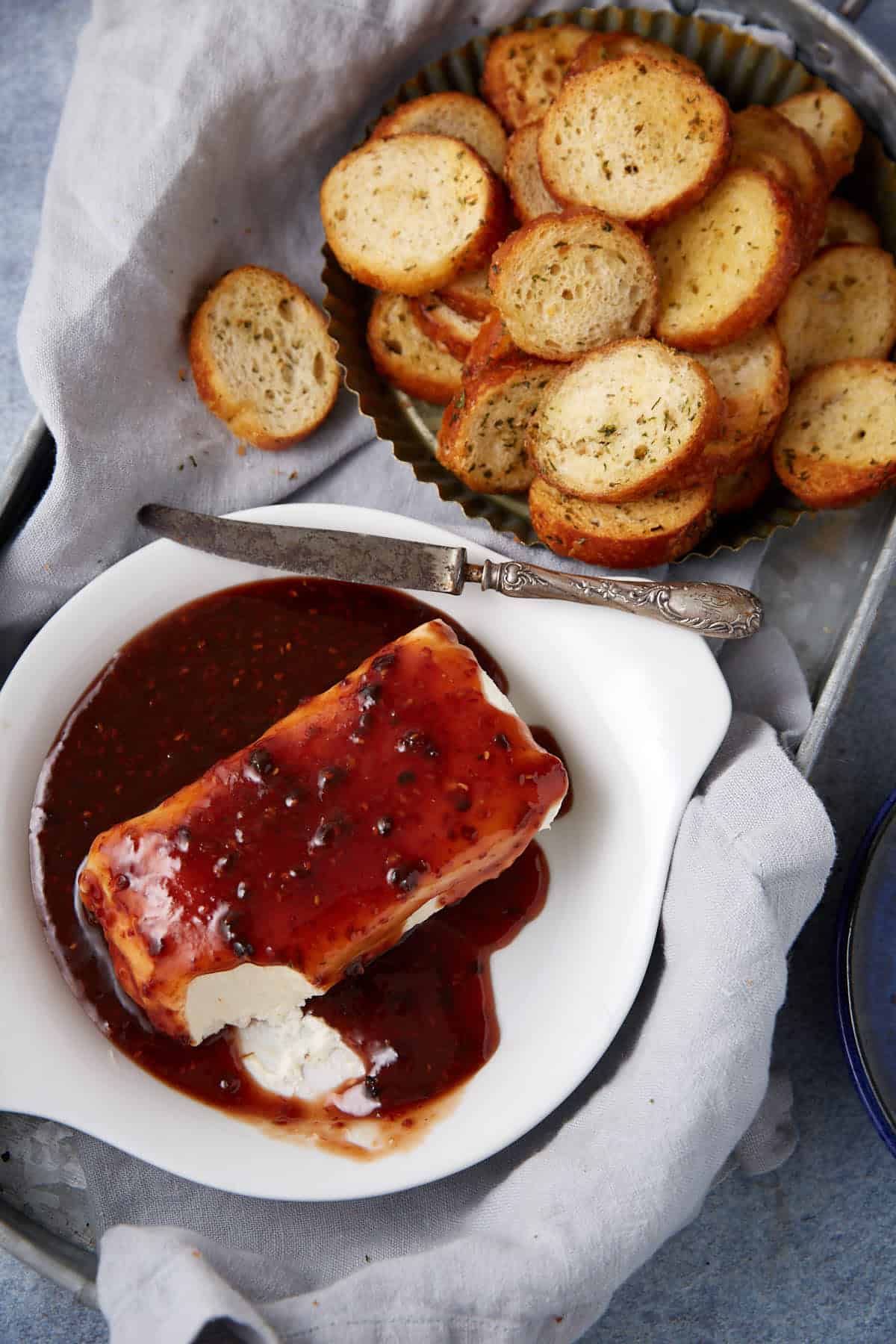 A white dish with a block of goat cheese covered in dark berry sauce sits next to a knife. Behind it, a tray is filled with round toasted bread slices, perfect for enjoying with this raspberry cream cheese dip.
