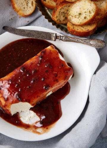 A block of cream cheese topped with pepper jelly sits on a white dish, served with a spreading knife and slices of toasted bread—perfect for enjoying as a raspberry cream cheese dip appetizer.