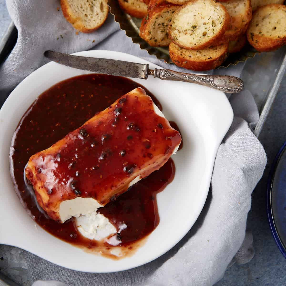 A block of cream cheese topped with pepper jelly sits on a white dish, served with a spreading knife and slices of toasted bread—perfect for enjoying as a raspberry cream cheese dip appetizer.