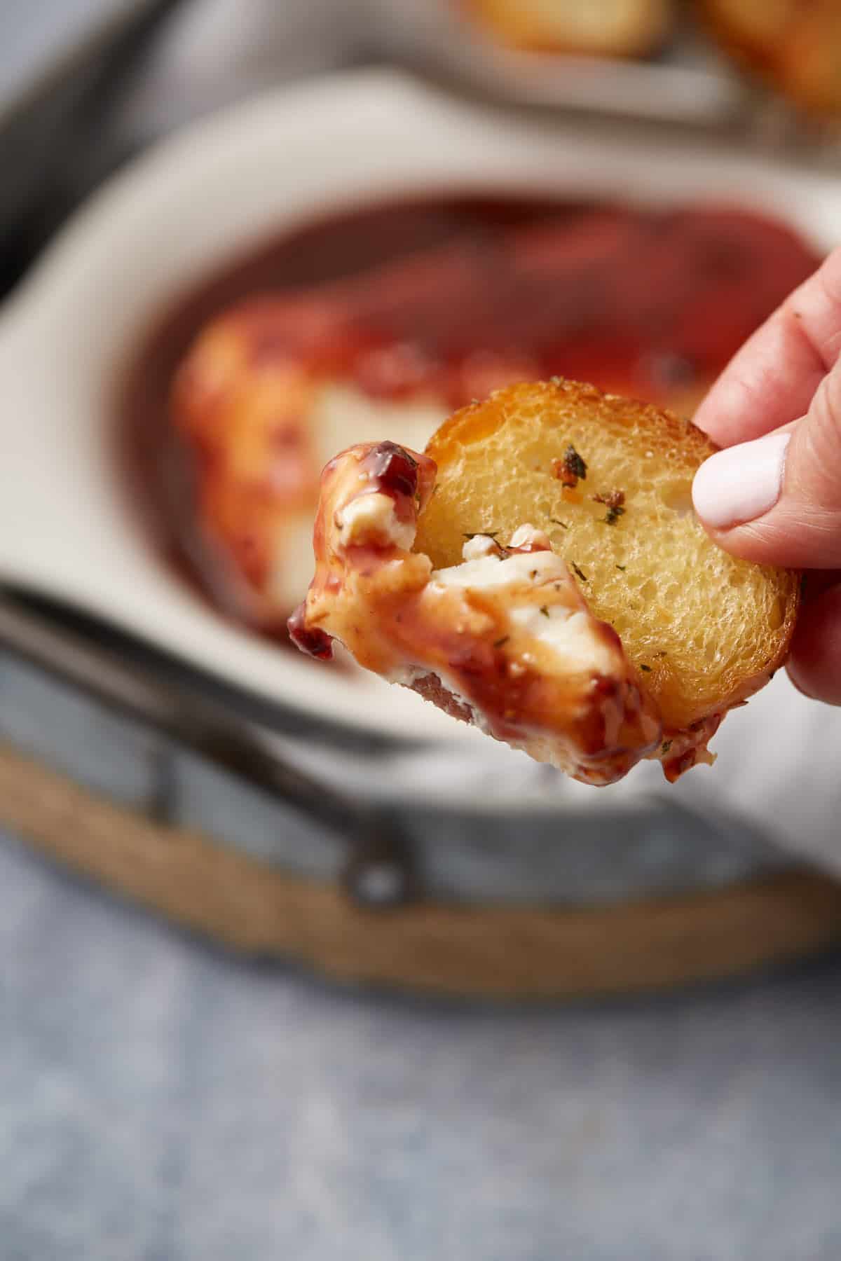 A hand holds a toasted bread slice topped with melted cheese and tomato sauce, with a dish of baked cheese, sauce, and raspberry cream cheese dip in the background.