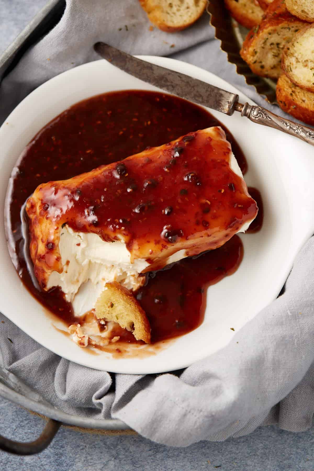 A block of raspberry cream cheese dip topped with raspberry sauce sits on a white plate, surrounded by a knife and toasted bread slices. A piece of bread is dipped into the creamy spread. A gray napkin lines the tray underneath.