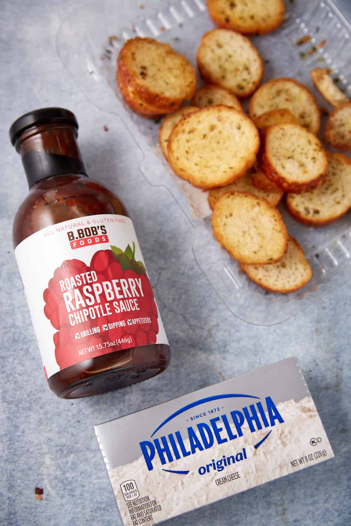 A bottle of roasted raspberry chipotle sauce, a package of Philadelphia original cream cheese, and a container of toasted baguette slices await to create a delicious raspberry cream cheese dip on a light gray surface.