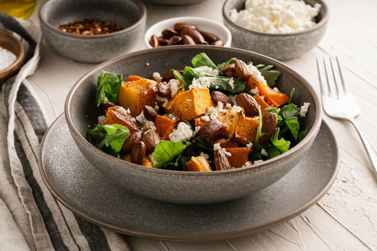 A bowl of salad with roasted pumpkin, leafy greens, crumbled cheese, and almonds, surrounded by small bowls of nuts, cheese, and spices—perfect for homestyle fall meals that taste like home, served on a light wooden table with a striped cloth and fork.