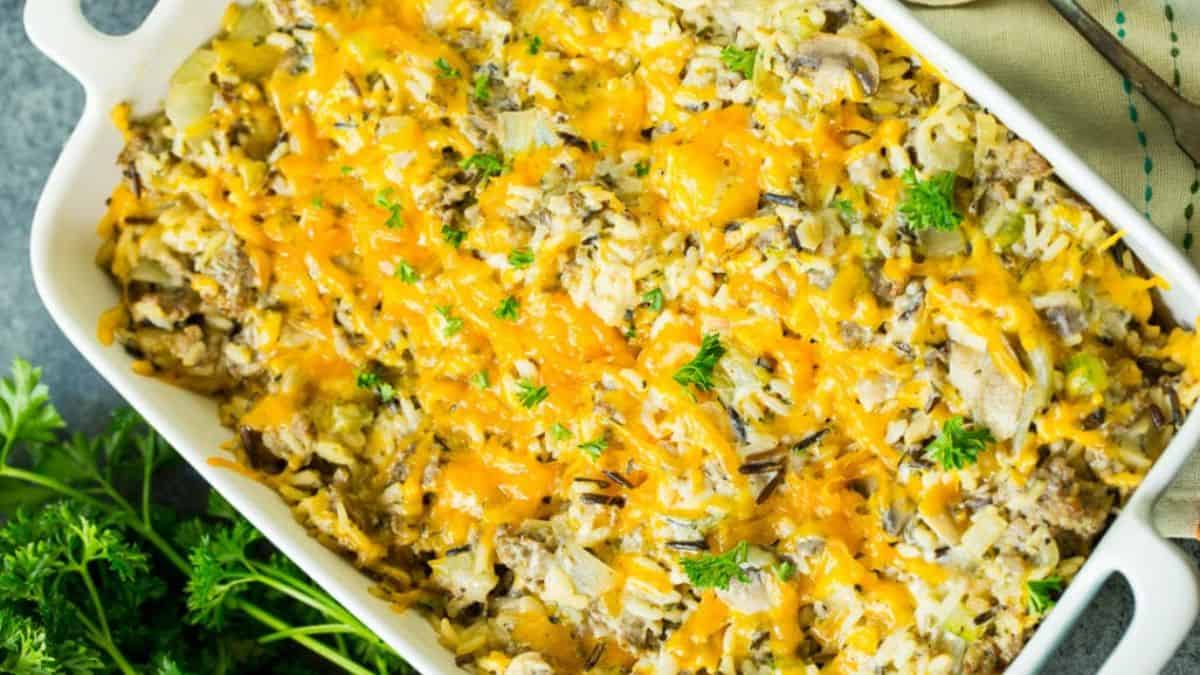 A casserole dish filled with rice, mushrooms, and ground meat, topped with melted cheddar cheese and garnished with fresh parsley. A bunch of parsley is beside the dish on a gray surface.