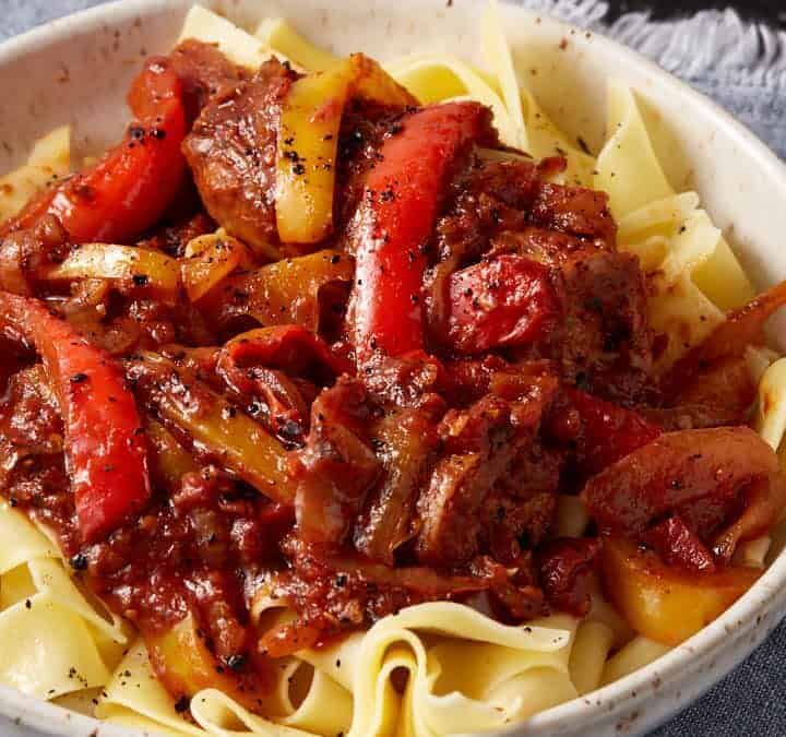 A bowl of wide egg noodles topped with a rich beef and bell pepper stew, garnished with black pepper, sits on a blue cloth napkin.