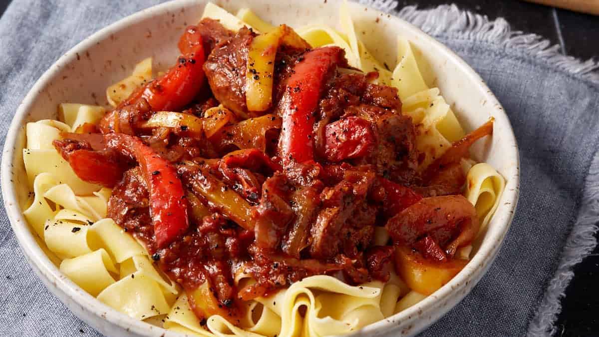 A bowl of wide egg noodles topped with a rich beef and bell pepper stew, garnished with black pepper, sits on a blue cloth napkin.