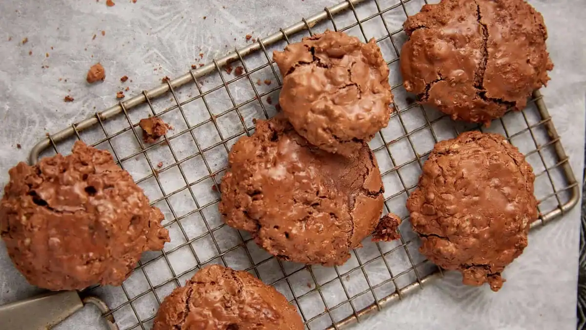 Six chocolate cookies with a cracked, glossy surface rest on a metal cooling rack lined with parchment paper. The cookies look chunky and rich, with a slightly rustic appearance.