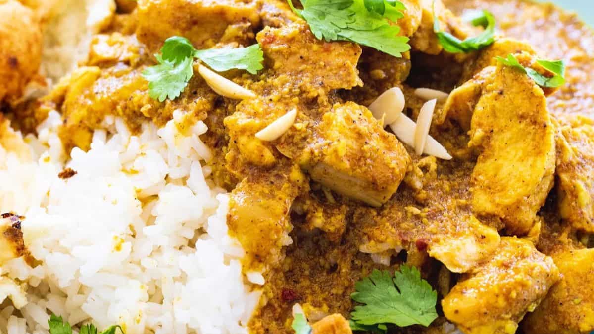 Close-up of a plate with white rice topped with a yellow-brown curry, chunks of chicken, fresh cilantro leaves, and slivered almonds.