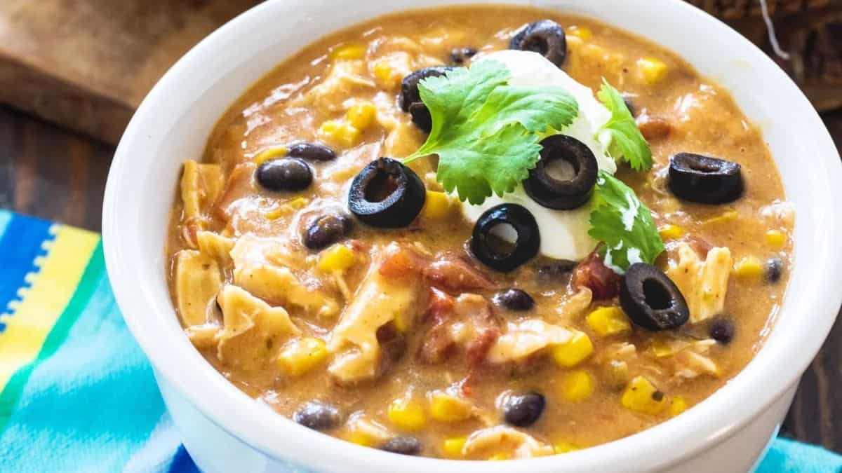 A bowl of creamy chicken enchilada soup topped with sliced black olives, a dollop of sour cream, and fresh cilantro, with visible corn, beans, and tomato pieces. A colorful napkin is beside the bowl.