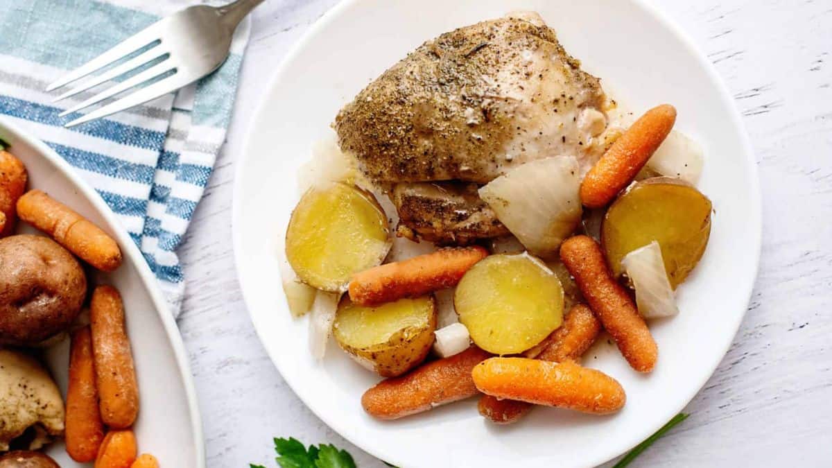 A white plate with baked chicken thigh, sliced baby potatoes, baby carrots, and cooked onions, next to a fork and a dish of more vegetables on a striped towel.