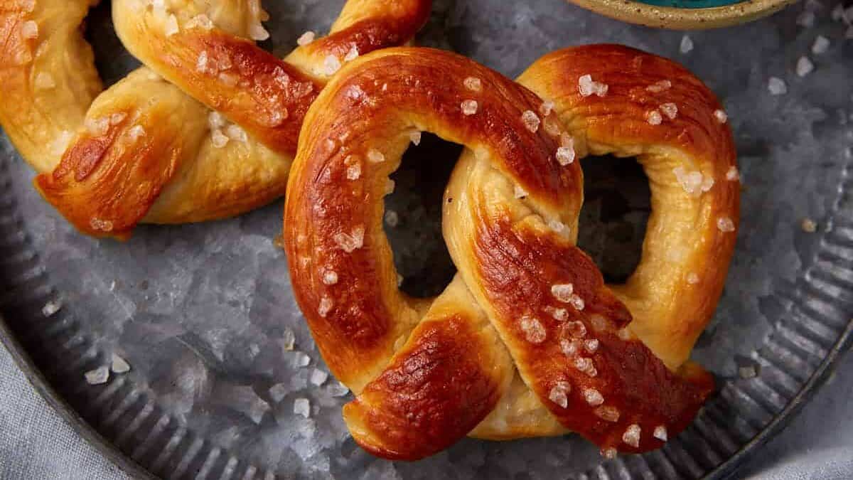 Two soft pretzels sprinkled with salt rest on a round metal tray beside a bowl of creamy dip topped with black seeds, a knife placed on the bowl. The tray sits atop a gray cloth.