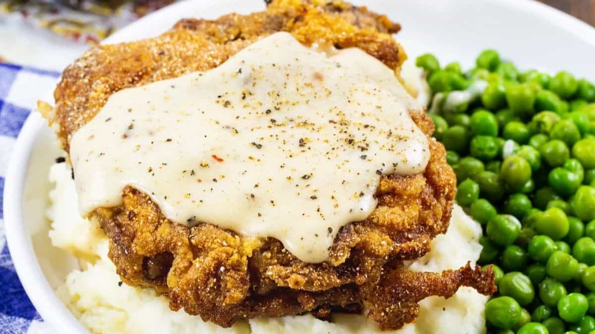 A serving of chicken fried steak topped with creamy peppered gravy sits on mashed potatoes, with a side of green peas on a white plate.