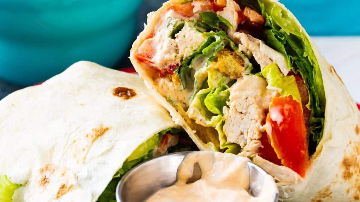 A close-up of a chicken wrap filled with lettuce, tomato, and sauce, cut in half, served next to a small metal cup of creamy dipping sauce.
