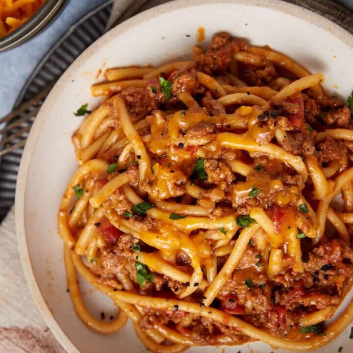 A plate of easy taco spaghetti—thick noodles topped with seasoned ground meat, tomato sauce, and melted cheddar cheese, garnished with herbs—sits on a cloth napkin with a fork nearby.