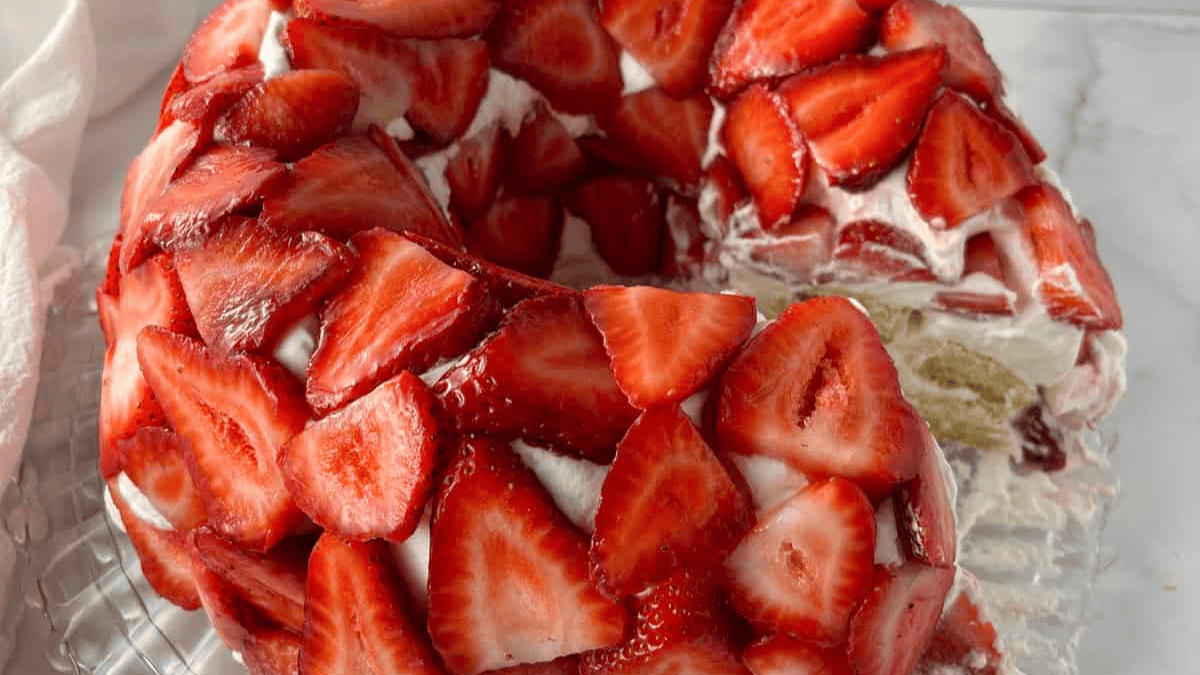 A bundt cake covered with layers of whipped cream and fresh, sliced strawberries, displayed on a glass plate with a slice missing to show the cake’s interior.