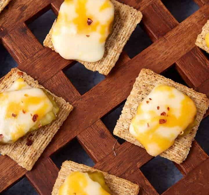 A lattice-style wooden tray holds a Dill Pickle Triscuit Appetizer—square wheat crackers topped with melted yellow and white cheese; some are finished with pickle slices and red pepper flakes.