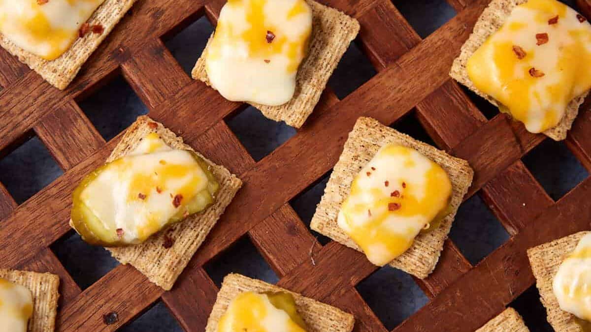A lattice-style wooden tray holds a Dill Pickle Triscuit Appetizer—square wheat crackers topped with melted yellow and white cheese; some are finished with pickle slices and red pepper flakes.
