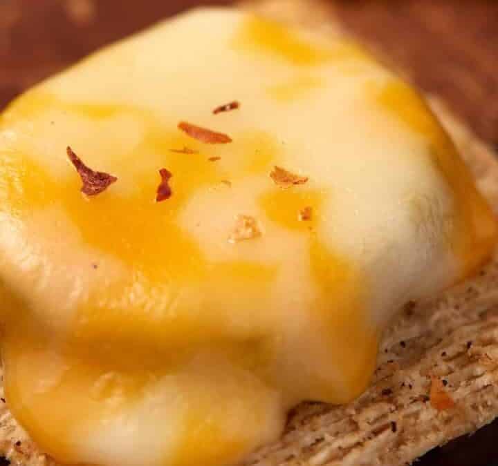 A Dill Pickle Triscuit Appetizer features a square cracker topped with melted cheddar and mozzarella cheese, sprinkled with red pepper flakes, and served on a wooden surface.