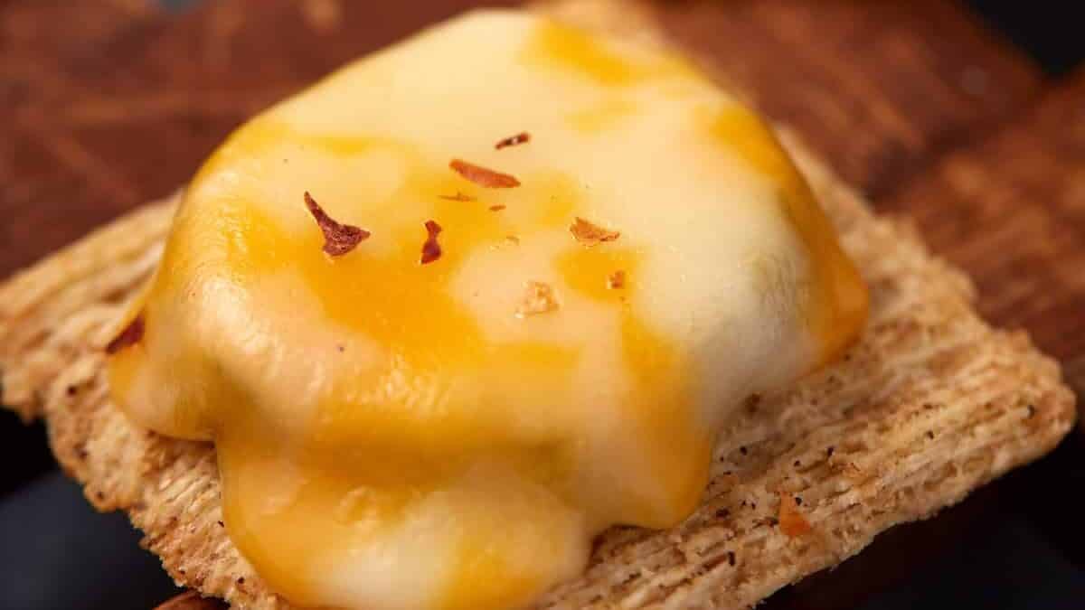 A Dill Pickle Triscuit Appetizer features a square cracker topped with melted cheddar and mozzarella cheese, sprinkled with red pepper flakes, and served on a wooden surface.