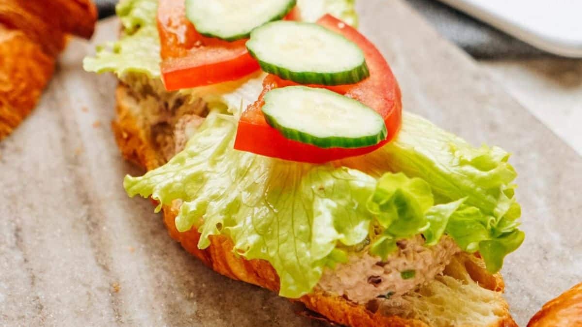 A croissant sandwich topped with leafy lettuce, sliced tomato, and cucumber, filled with a creamy tuna salad—a classic example of things Americans call salad that aren't salad—displayed on a light-colored surface.