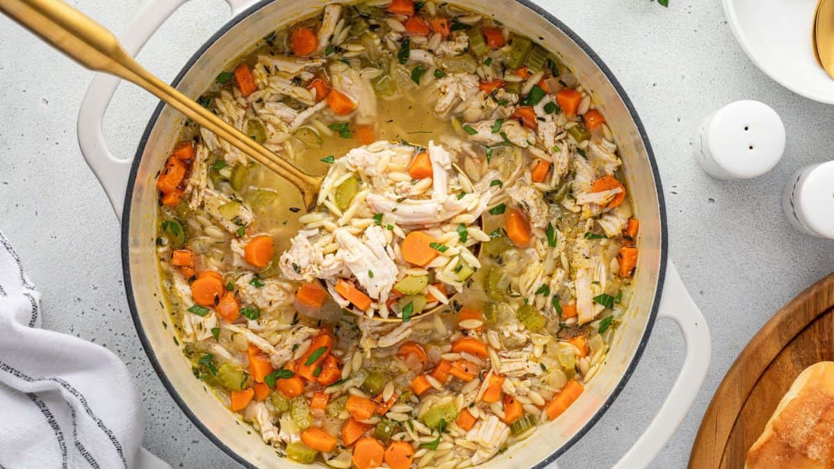 A pot of chicken orzo soup with shredded chicken, carrots, celery, and herbs, being stirred with a gold ladle. The soup is hearty and colorful, with visible chunks of vegetables and pasta.