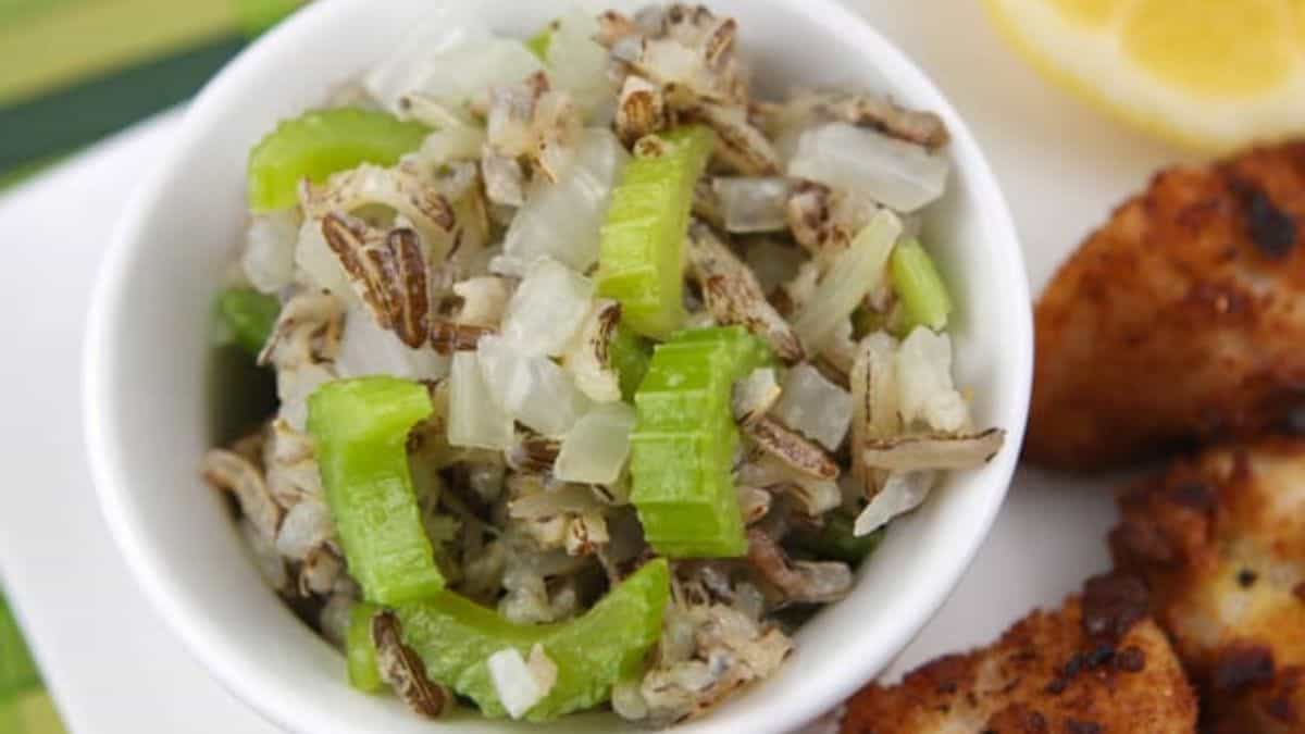 A white bowl filled with wild rice, diced onions, and celery slices, with a plate of seasoned, cooked meat and a lemon wedge partially visible beside it.