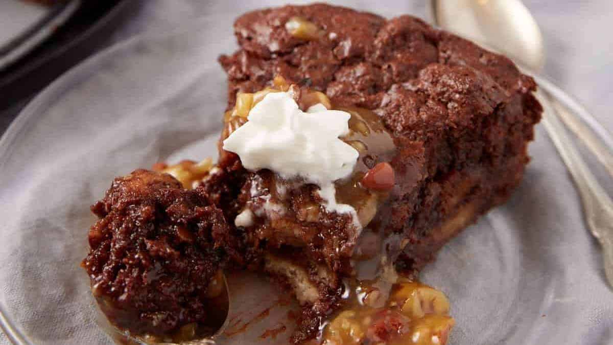 A slice of rich chocolate cake topped with caramel sauce, chopped nuts, and a dollop of whipped cream on a glass plate, with a spoon beside it.