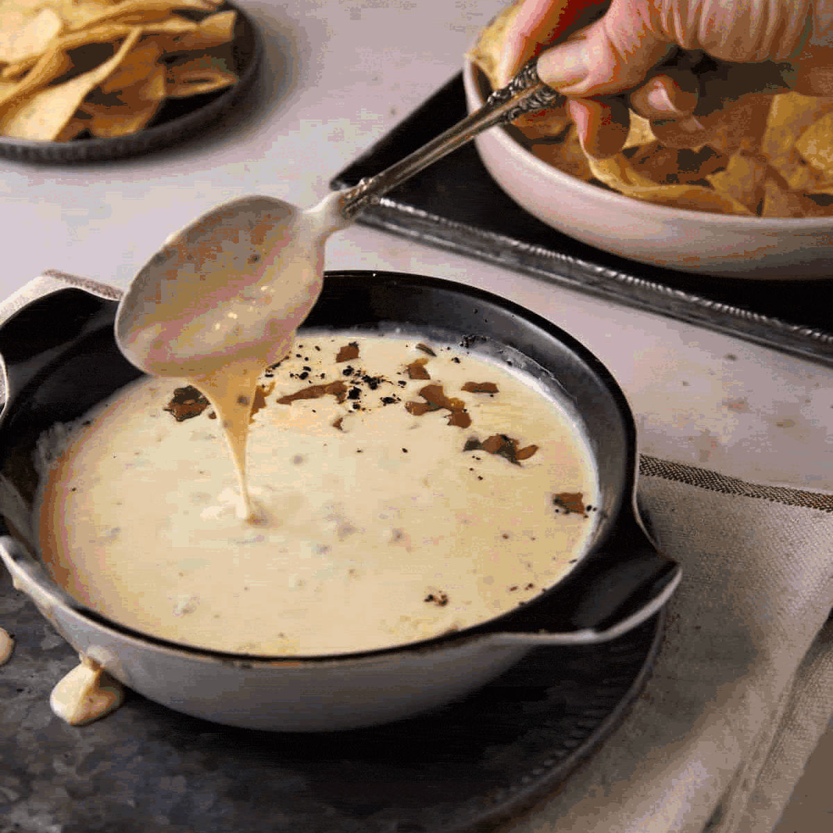A hand holds a spoon above a black bowl filled with creamy cheese dip, with some dip dripping from the spoon. Crushed peppers garnish the dip. Bowls of tortilla chips are visible in the background.